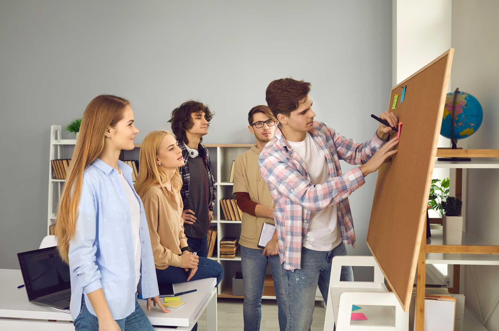 Young students are brainstorming on a board. One young man is writing on a sticky note while his fellow students watch.