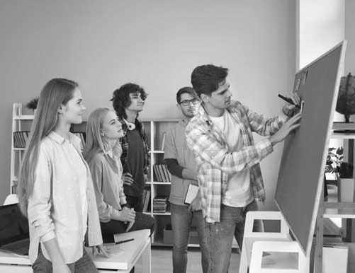 Young students are brainstorming on a board. One young man is writing on a sticky note while his fellow students watch.